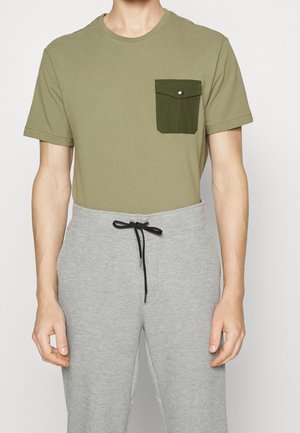 Mann trägt olivgrünes T-Shirt mit dunkelgrüner Brusttasche mit Knöpfen und hellgraue Jogginghose mit schwarzer Kordel.