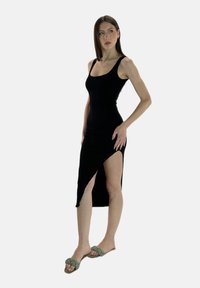 Abito nero senza maniche con design aderente e spacco laterale, realizzato in un materiale liscio. Abbinato a sandali piatti intrecciati verdi.