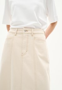 Beige Denimrock mit Taschen vorne, sichtbaren Nähten und einem Knopfverschluss vorne, kombiniert mit einem weißen, kurzärmligen Oberteil.