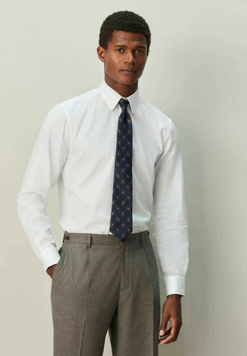 Bela srajca z vzorcem teksture, združena s sivimi hlačami. Modri kravata ima geometrični vzorec z zlatimi poudarki.