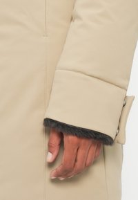 Beige puffer coat mouw met een zachte, donkere bontvoering. Kenmerkt zich door een gladde, gewatteerde textuur en een knoopdetail bij de manchetten.