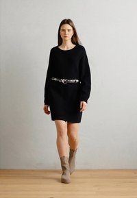 Svart strikket kjole med lange ermer og en avslappet passform, belte i midjen med et sølvbelte. Modellen har på seg beige ankelstøvletter.