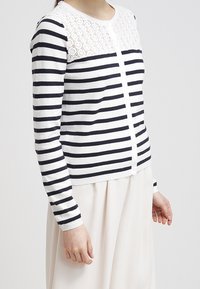 Pruhovaný kardigan s navy a bílými vodorovnými pruhy, krajkovými detaily na ramenou a knoflíkovým zapínáním vpředu. Měkká textura.