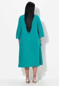 Vestito teal fino al ginocchio realizzato in tessuto leggero, dotato di maniche a tre quarti e un taglio dritto. Sandali con tacco argento completano il look.