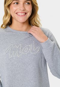 Sivá mikina s dlhými rukávmi, s dizajnom "Moi" vytvoreným z malých bielych perál. Mäkká, textúrovaná tkanina s voľným strihom.