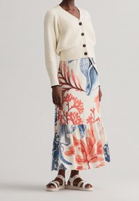 Cardigan lavorato a maglia color crema con tre bottoni marroni, abbinato a una gonna lunga floreale con disegni blu e corallo. Sandali bianchi con cinturini.