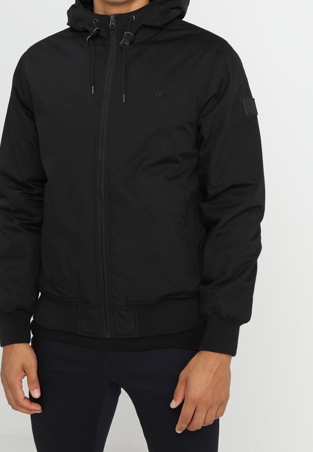 Homme portant une veste à capuche noire zippée avec des poignets et un ourlet côtelés, debout devant un fond clair uni.