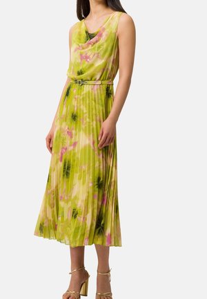 Frau trägt ärmelloses, plissiertes Midikleid mit grünem, pinkem und schwarzem Blumenmuster und dünnem Gürtel, kombiniert mit goldenen Riemchenabsätzen.