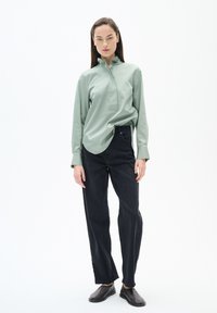 Hellgrünes Langarmhemd mit Kragen und Knopf am Hals, kombiniert mit dunklen, geraden Jeans und dunklen Schuhen.