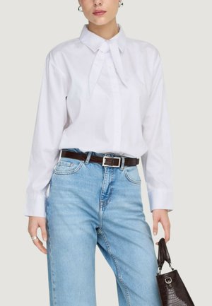 Femme portant un chemisier blanc à manches longues avec un détail unique au niveau de la cravate, un jean bleu clair avec une ceinture noire, tenant un sac à main marron foncé.