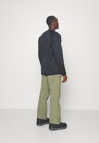 Chemise noire à manches longues avec encolure ronde, associée à un pantalon cargo vert olive avec un logo sur le côté et des bottes noires imperméables.