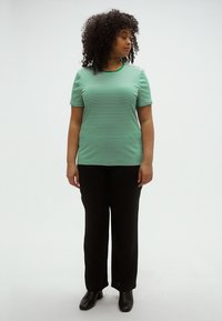 Žaliai ir baltai dryžuota trumpomis rankovėmis marškinėliai, derinami su juodomis plačiomis kelnėmis ir juodais kulkšnies batais, fone yra balta sienos.