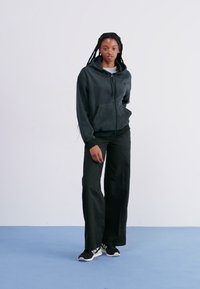 Černá mikina na zip s měkkou texturou, kombinovaná s volnými černými kalhotami a černými teniskami. Model stojí na světlém pozadí.