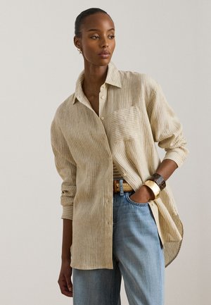 Γυναίκα που φοράει μπεζ ριγέ πουκάμισο με κουμπιά, μπλε τζιν, χρυσά και καμηλό χρωματιστά βραχιόλια, στέκεται με το ένα χέρι στην τσέπη.