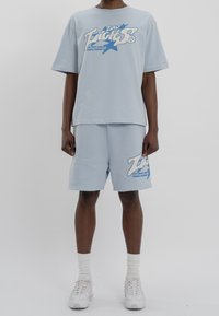 Světle modré bavlněné tričko a šortky s grafickým potiskem. Volný střih, kulatý výstřih a žebrované manžety. Bílé sportovní boty a ponožky.