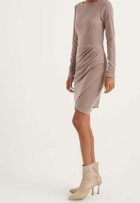 Abito a maniche lunghe in rib knit color taupe con dettagli drappeggiati sul lato. Abbinato a stivali aderenti beige chiaro con tacco a stiletto.