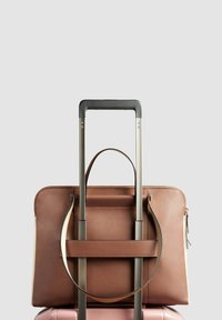 Braune Lederhandtasche mit strukturiertem Design, ausgestattet mit oberen Griffen und einer Fronttasche, die auf einem pinken Koffergriff ruht.