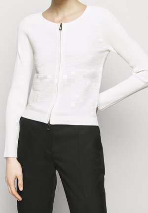Femme portant un haut blanc côtelé à manches longues avec fermeture éclair, associé à un pantalon noir taille haute, posant avec une main sur la hanche.