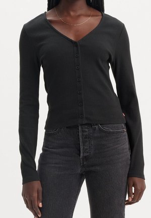 Femme portant un haut côtelé noir ajusté à manches longues avec boutons et un jean gris foncé avec un fin collier en or.
