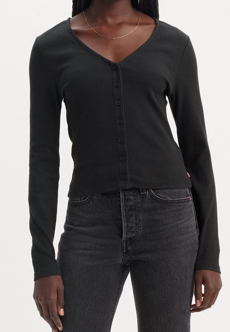 Femme portant un haut côtelé noir ajusté à manches longues avec boutons et un jean gris foncé avec un fin collier en or.