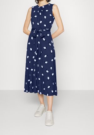 Ærmeløs navyblå midi-kjole med hvide uregelmæssige polkaprikker, bånd til at binde i taljen, båret med hvide sneakers, model stående med hænderne bag ryggen.