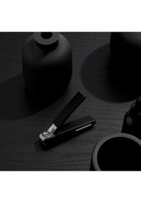 Zwarte Tweezerman nagelknipper op een donkerhouten oppervlak, omringd door matzwarte afgeronde potjes en een pipetflesje.