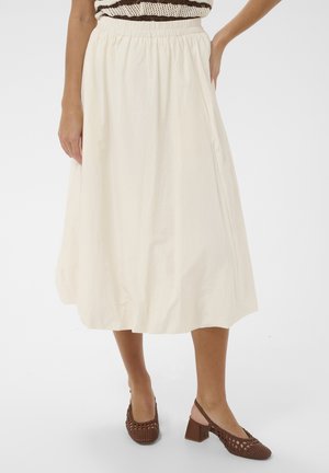 Kobieta nosi kremową spódnicę midi z elastycznym pasem oraz brązowe plecione sandały na słupku z odkrytą piętą, stoi na białym tle.