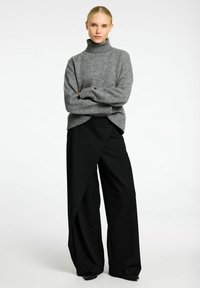 Pull en maille gris avec col roulé et poignets côtelés, associé à un pantalon large noir à coupes droites avec un design angulaire et drapé.
