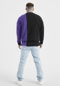 En man med nacktatuerng står vänd bort, klädd i en sweatshirt som är hälften lila och hälften svart, ljusblå jeans med vit logotryck och vita sneakers.