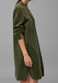 Donna che indossa un vestito in velluto a coste verde oliva con maniche lunghe e bottoni sul davanti, con le maniche rimboccate, su uno sfondo grigio.