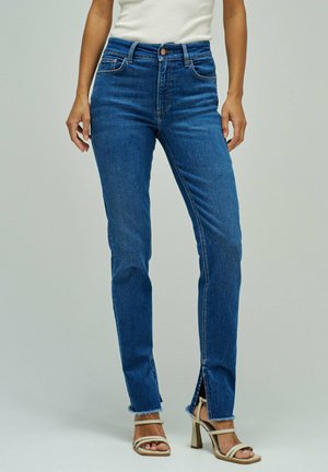 Mujer con vaqueros de talle alto azules con aberturas frontales en las piernas y dobladillos sin rematar, combinados con sandalias beige de tacón alto con tiras y una camiseta blanca sin mangas.