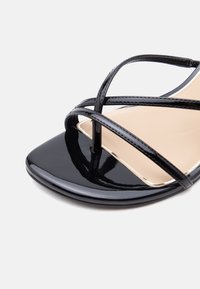 Μαύρο σανδάλι από δερματίνη με λεπτά, σταυρωτά λουριά και τετράγωνη μύτη σε ανοιχτό φόντο.