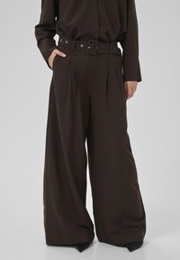 Tummanruskeat pinstripe-malliset leveät housut vyöllä, kantavat samanlaista paitaa, käsi taskussa, seisoo yksinkertaista taustaa vasten.