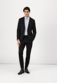 Schwarze maßgeschneiderte Sakko über einem hellblauen Hemd, kombiniert mit schwarzen Slim-Fit-Hosen und glänzenden schwarzen Stiefeletten. Schlichter Hintergrund.