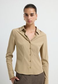 Camicia lunghe maniche beige chiaro con colletto a punta, chiusura con bottoni e un tessuto leggermente strutturato. Vestibilità su misura con orlo curvo.