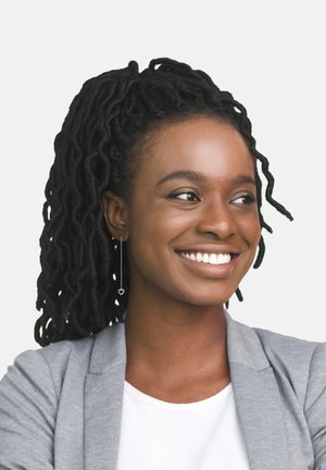 Persona sorridente con lunghe trecce nere, che indossa un blazer grigio chiaro sopra una camicia bianca e un orecchino a forma di cuore.