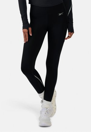 Persona vistiendo mallas negras de Reebok con rayas reflectantes, calcetines blancos y zapatillas blancas de correr sobre un fondo liso.