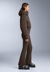 Ruskea eristetty takki korkealla kauluksella ja etutaskuilla, yhdistetty istuviin ruskeisiin housuihin. Mustat saappaat hopein koristeyksityiskohdin.