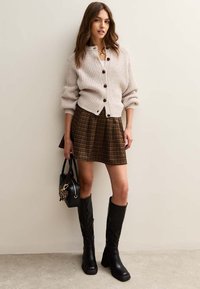 Cardigan beige a coste con bottoni marroni, abbinato a una gonna a scacchi marrone e stivali neri con tacco alti fino al ginocchio, completato da una borsa nera.