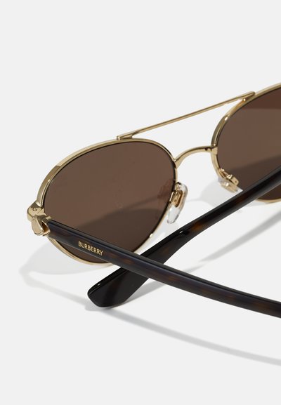Nahaufnahme von Sonnenbrillen mit goldfarbenem Rahmen, braunen Gläsern und schwarzen Bügeln, die das Burberry-Logo am Bügel zeigen.
