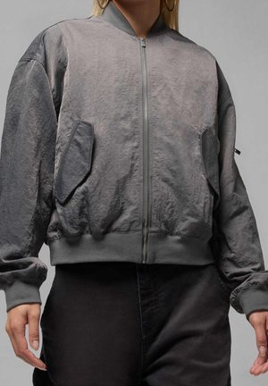 Personne portant une veste bomber grise zippée avec de grandes poches à rabat et des poignets côtelés, associée à un pantalon noir sur un fond uni.