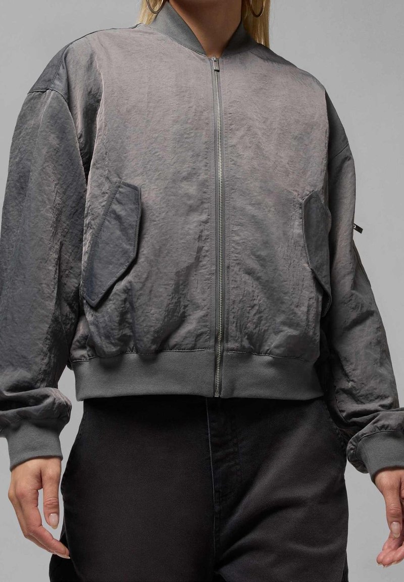 Personne portant une veste bomber grise zippée avec de grandes poches à rabat et des poignets côtelés, associée à un pantalon noir sur un fond uni.