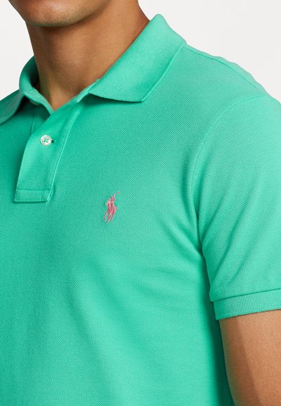 Polo turquoise en tissutexturé, avec patte de boutonnage à trois boutons et un petit logo rose sur la poitrine gauche. Manches courtes.