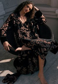 Lunga veste nera con motivi floreali in bronzo, indossata sopra un abito coordinato, seduta su una sedia testurizzata, scalza su una superficie in pietra.