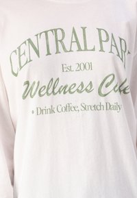 Bijela majica dugih rukava s zelenim natpisom "CENTRAL PARK Est. 2001 Wellness Cafe Pij kavu, isteži se svakodnevno."