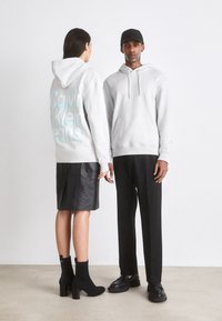 I felpe grigie presentano un audace logo "Calvin Klein Jeans" sulla schiena. I modelli indossano una gonna in pelle nera e pantaloni, abbinati a calzature nere.