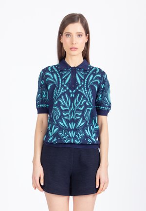 Kobieta ubrana w granatową dzianinową koszulkę polo z turkusowymi wzorami kwiatowymi i granatowe krótkie spodenki, stojąca na tle prostej białej ściany.