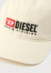 Μπεζ καπέλο μπέιζμπολ από ραβδωτό ύφασμα με κεντημένο κόκκινο και μαύρο λογότυπο Diesel και κείμενο "DENIM DIVISION" από κάτω.