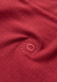 Tessuto lavorato a maglia rosso con una superficie testurizzata; presenta un logo circolare ricamato nell'area inferiore con un motivo decorativo di alloro.
