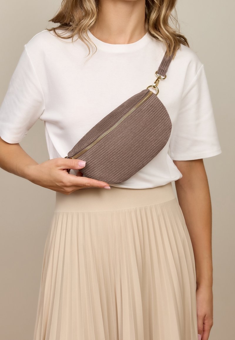 Femme portant un t-shirt blanc et une jupe plissée beige tient un sac bandoulière côtelé taupe avec une fermeture éclair dorée sur sa poitrine.